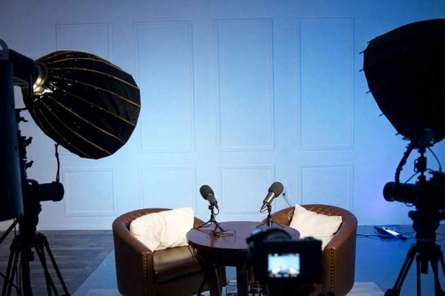 En podd-/intervjusetup med mikrofoner, fåtöljer och ljusutrustning mot en blå vägg.